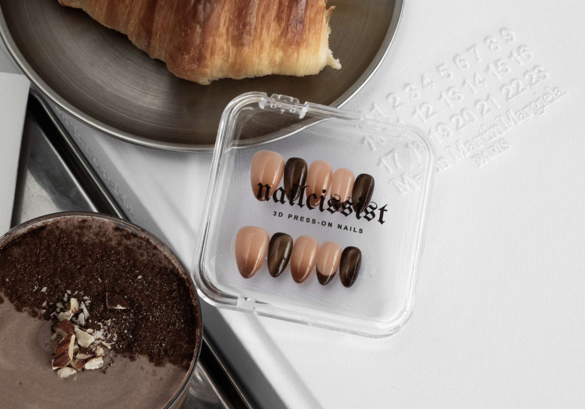 Set of press-on nails in a clear case on a white surface with a croissant and coffee.